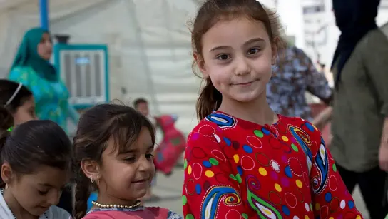 Ein kleines Mädchen in einem roten und bunt gemusterten Pulli lächelt in die Kamera. Neben ihr sind 3 weitere Kinder zu sehen.