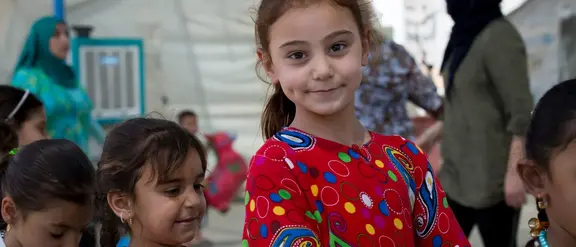 Ein kleines Mädchen in einem roten und bunt gemusterten Pulli lächelt in die Kamera. Neben ihr sind 3 weitere Kinder zu sehen.