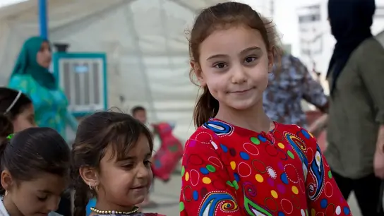 Ein kleines Mädchen in einem roten und bunt gemusterten Pulli lächelt in die Kamera. Neben ihr sind 3 weitere Kinder zu sehen.