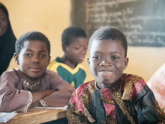 In der Schule: 3 Schwarze Kinder sitzen an Schulbänken. Sie haben kurzes Haar und grinsen in die Kamera. 