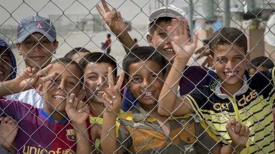 Eine Gruppe Kinder mit gebräunter Haut und dunklen Haaren stehen hinter einem Maschendrahtzaun und lachen und winken in die Kamera.