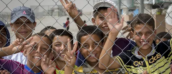 Eine Gruppe Kinder mit gebräunter Haut und dunklen Haaren stehen hinter einem Maschendrahtzaun und lachen und winken in die Kamera.