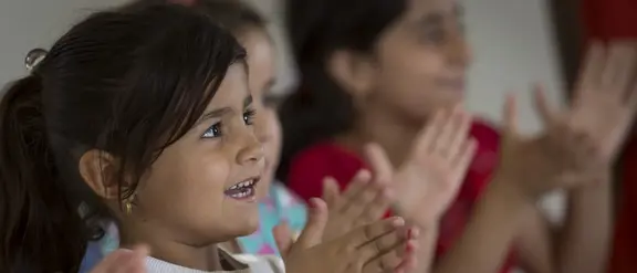 Ein Kind mit langem, dunklem Haar schaut auf etwas abseits der Kamera und lacht und klatscht. Hinter ihm sind verschwommen andere Kinder zu sehen, die das gleiche tun.