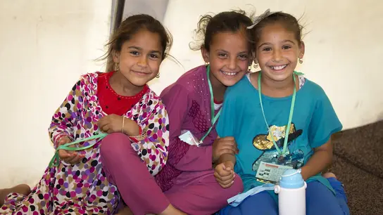 Drei Kinder mit dunkler Haut und bunten Kleidern sitzen auf dem Boden und lachen in die Kamera.